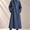 بارانی مینیمال با یقه دراپه مورب و تک‌دکمه چوبی شناور در پس‌زمینه بتنی بافت‌دار | Minimalist asymmetric drape trench coat with raglan sleeves and wooden accent button on textured neutral background