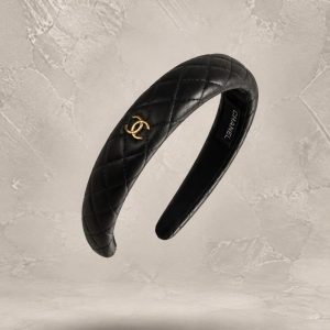 هدبند چرم طبیعی مشکی شنل با دوخت کوئلته ضربدری و لوگوی فلزی طلایی دو سر C در پس‌زمینه بافت‌دار سنگی | Black Chanel quilted leather headband with interlocking gold CC logo in a textured stone background