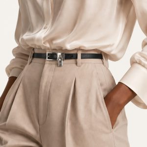 کمربند چرم مشکی ارمز با سگک فلزی نقره‌ای و آویز قفل کوچک روی پس‌زمینه بژ | Hermès black Epsom leather belt with palladium buckle and miniature padlock charm on beige background